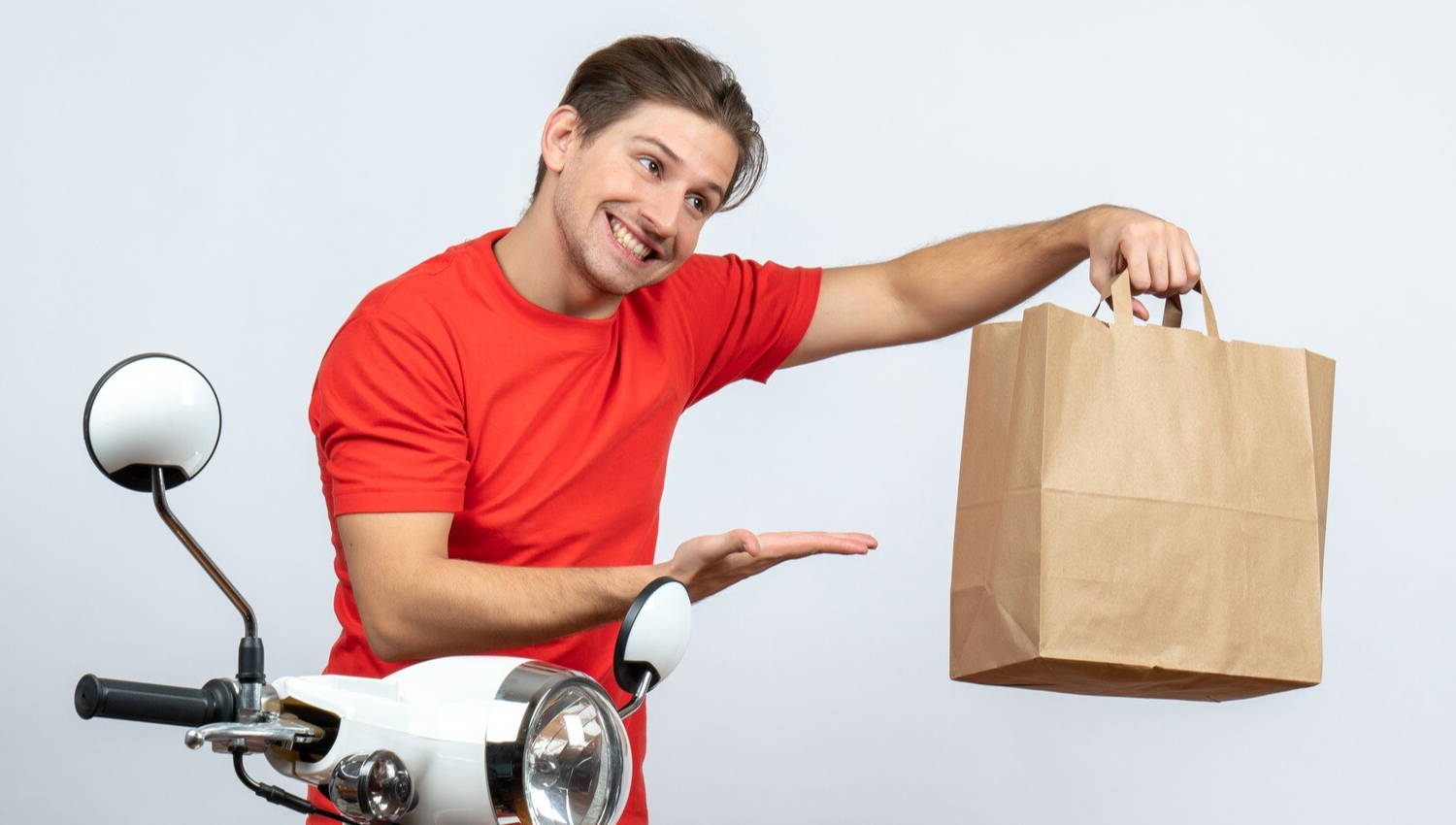 Smiling delivery man in red uniform standing near scooter pointing paper box on white background