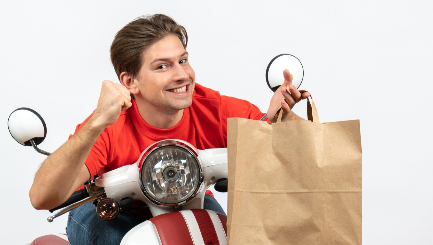 Young smiling proud courier guy in red uniform sitting on scooter holding paper bag on white wall