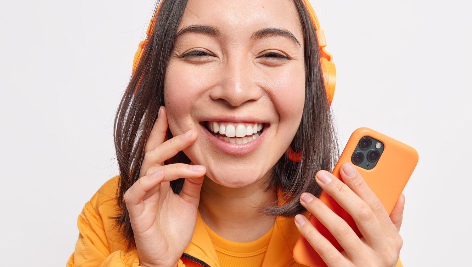 Close up shot of beautiful cheerful Asian woman smiles broadly from delight enjoys listening favorite music holds mobile phone dressed in orange jacket isolated over white wall.